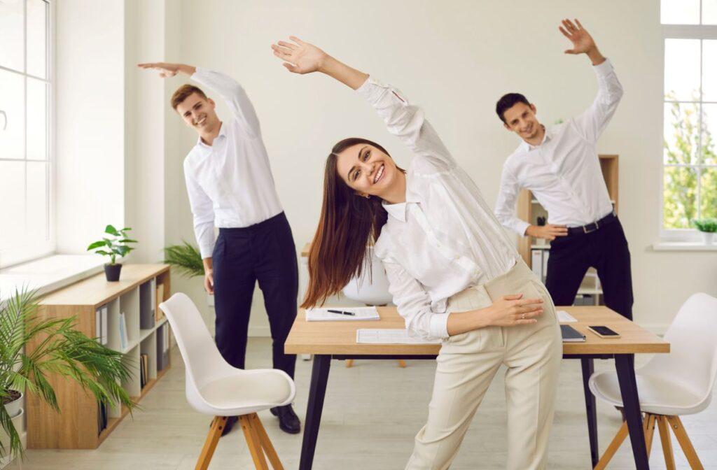 Drei Büroangestellte in Business-Kleidung machen Dehnübungen am Arbeitsplatz als Teil der betrieblichen Gesundheitsförderung.