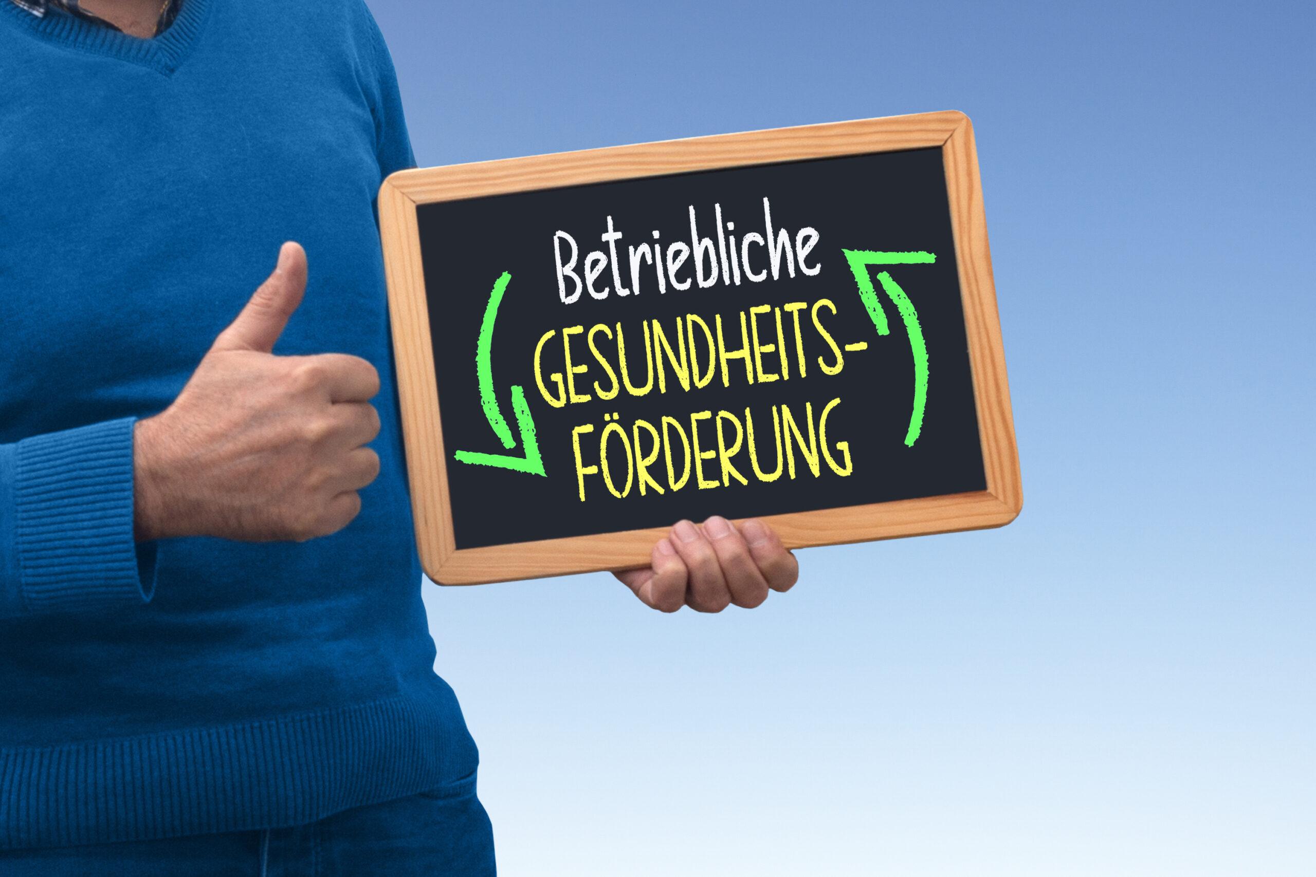 Person hält eine Tafel mit der Aufschrift „Betriebliche Gesundheitsförderung“ und zeigt mit einem Daumen hoch die Bedeutung des Themas.